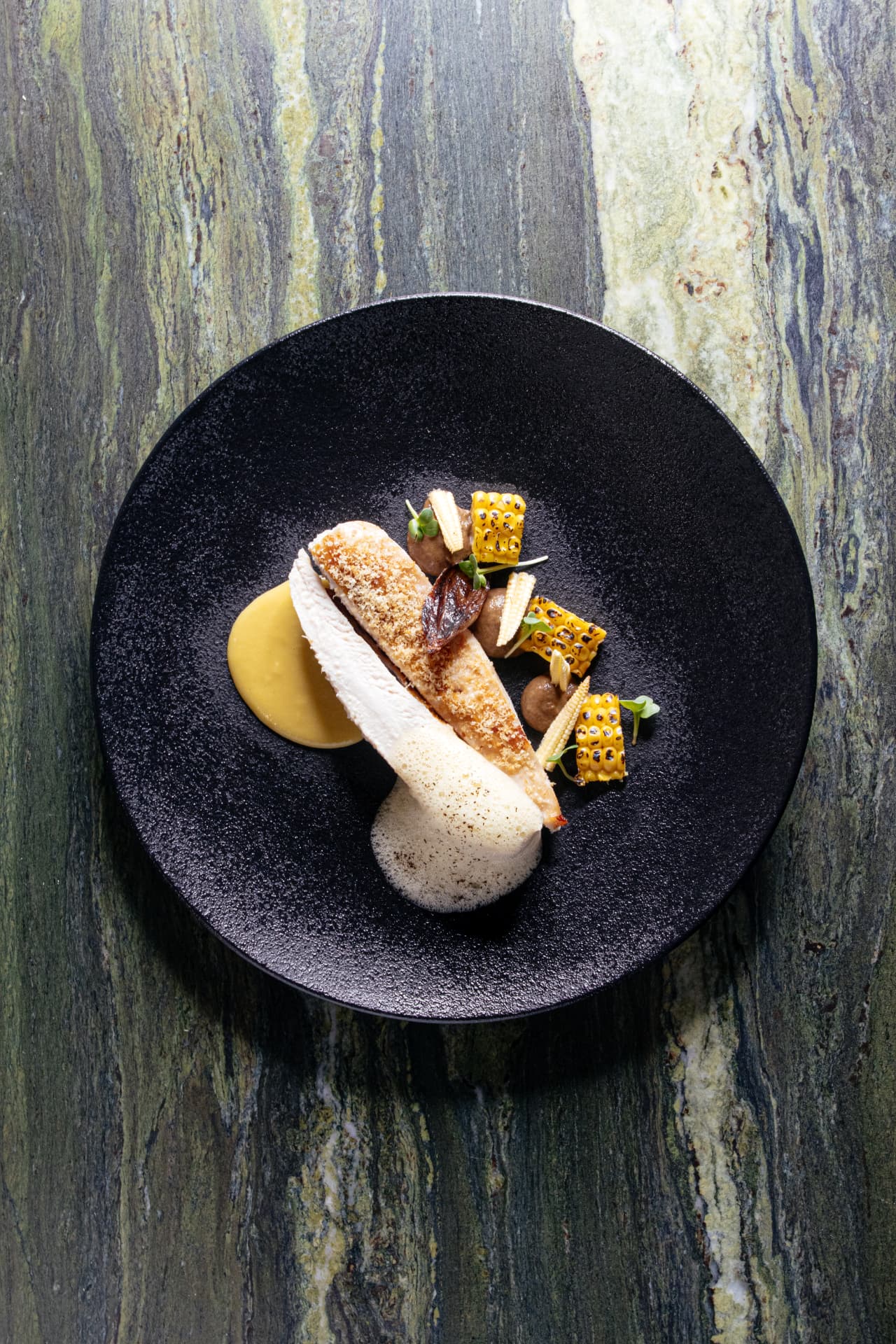 Poached chicken breast with grilled corn and foam on a black plate