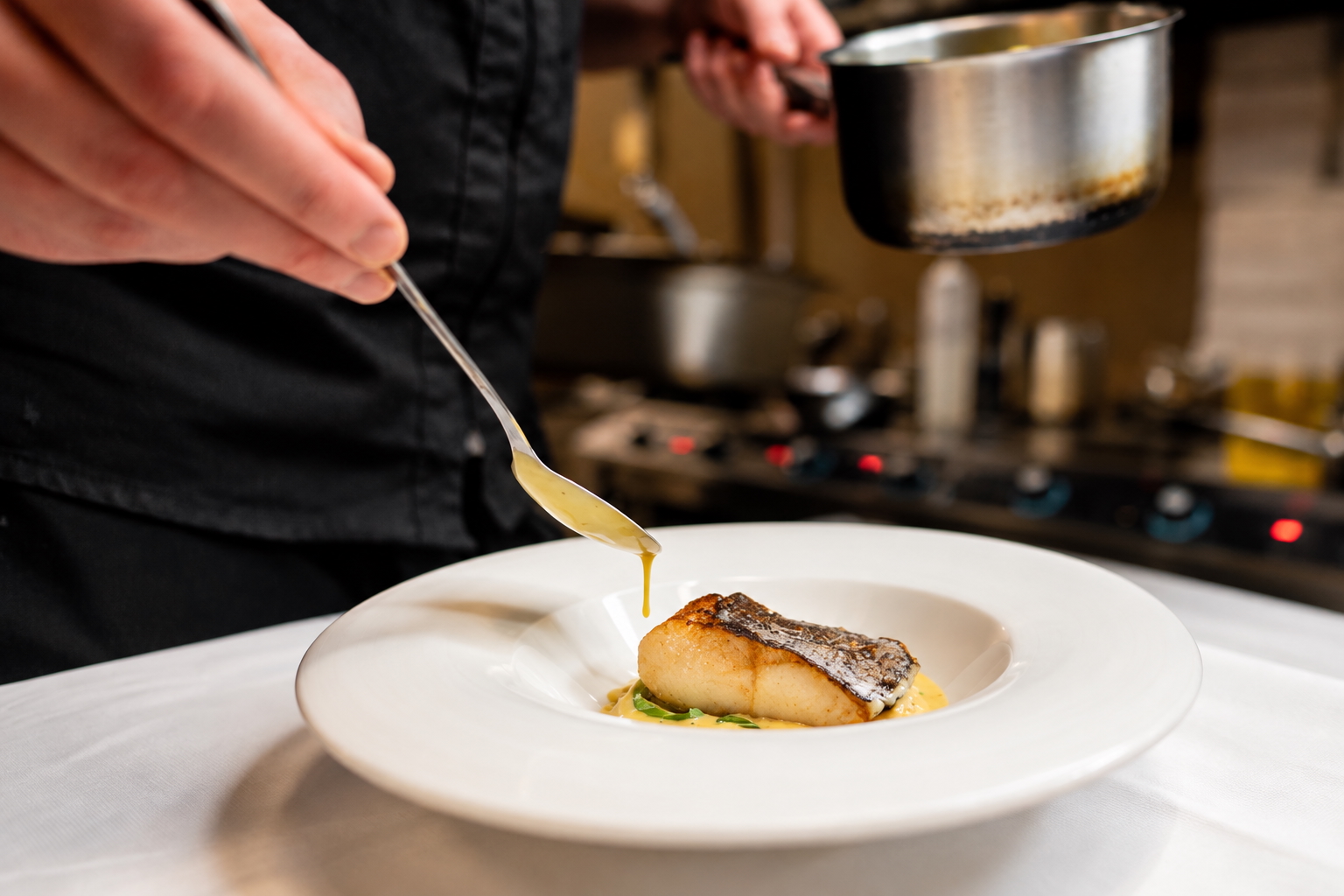 Chef's hands spooning sauce over a seared fish fillet in a white bowl